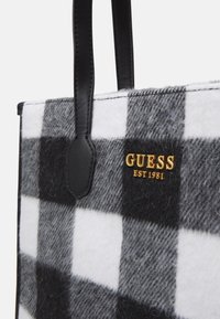 Sac fourre-tout en laine à carreaux noir et blanc, avec des poignées en cuir noir et un logo doré "GUESS EST 1981".