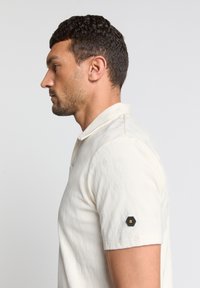 Korte mouwen polo shirt in roomkleur met een gestructureerd ruitpatroon. Heeft een klassieke kraag en een zwart hexagonaal logo patch op de linkermouw.