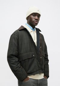 Mann in einer weißen Strickmütze, dunkler Jacke mit Vorderentaschen, blauen Hemd, cremefarbenem Reißverschluss-Pullover und grauen Jeans, mit Händen in den Taschen.