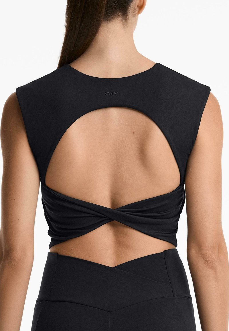 Top atletico nero con scollatura alta, schiena aperta e dettaglio attorcigliato. Realizzato in tessuto liscio, si abbina a pantaloni neri a vita alta.