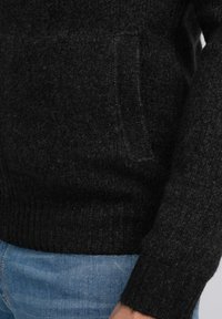 Zwarte gebreide trui met een gestructureerd oppervlak, geribbelde mouwen en een klein voorvakje. Draagt over lichtblauwe jeans.