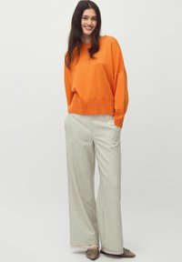 Maglione oversized arancione con spalle cadenti, abbinato a pantaloni larghi grigi chiari e scarpe flat beige, su uno sfondo semplice.