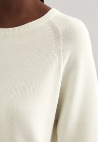 Pull en tricot crème avec un large col bateau, une finition texturée et des détails de couture visibles le long des épaules et des côtés.