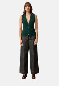 Zielona dziergana kamizelka z głębokim dekoltem w kształcie litery V, z zapięciem na guziki i dwiema kieszeniami z przodu, zestawiona z ciemnymi spodniami dżinsowymi z szerokimi nogawkami oraz brązowymi, masywnymi obcasami.
