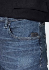 Blå jeans i denim med en klassisk rak passform. Har en framfick, synlig sömnad och en svart logotyp-patch på bakfickan.