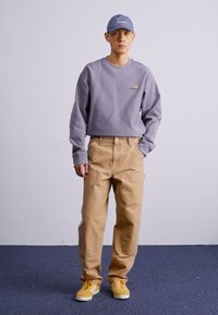 Sweatshirt lilas avec un logo, pantalon beige avec poches, baskets jaunes, et une casquette assortie, sur un fond uni.