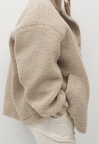 Beige fleecetrøje med en tekstureret overflade, der har en høj krave og oversized pasform. Syningerne er synlige langs sømmene.