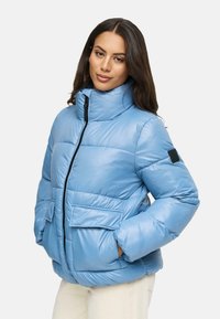 Ljusblåt puffjacka med hög krage, svart dragkedja, två framfickor och texturerad tyg. Mjuk, vadderad design med en accentpatch.