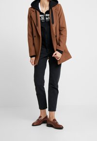 Abrigo de mezcla de lana marrón de doble botonadura sobre una sudadera negra con capucha, combinado con jeans de mezclilla oscuros y mocasines marrones con acentos metálicos.