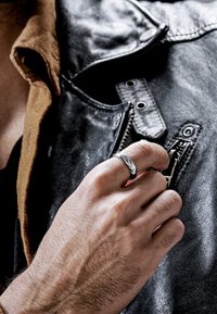 Sølvring med en teksturert, vridd design som bæres på hånden mot en svart skinnjakke. Jakken har en brun krage og metallbeslag.