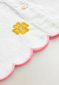 Fehér szövet, pink hullámos széllel és sárga hímzett virággal a közepén. Kiemelkedő rózsaszín kiegészítő gomb található rajta.