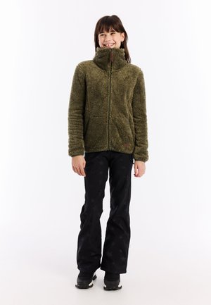Lachend meisje staat in een olijfgroen fleecejack, zwarte broek met sterrenpatronen en zwarte snowboots op een witte achtergrond.