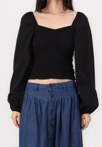 Haut court noir à encolure carrée et texture froncée. Manches longues bouffantes. Associé à une jupe en jean avec détails à boutons.