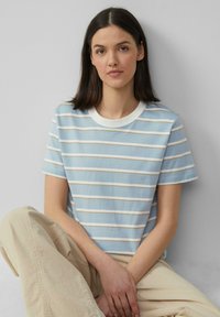 Lichtblauwe gestreepte t-shirt met korte mouwen, een witte ronde hals en horizontale strepen in beige. Model zit met beige broek.