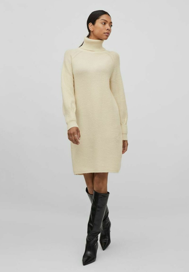 Robe pull en crème avec un col montant, des manches longues et une texture côtelée. Portée avec des bottes noires à talons aigus jusqu'aux genoux.
