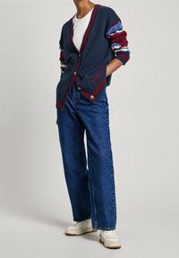Cardigan navy con maniche a strisce rosse e blu, chiusura a bottoni e dettaglio del logo. Abbinato a jeans blu a vita alta e sneakers bianche.