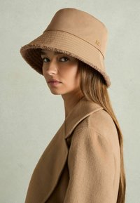 Brown bucket hat with a textured, fuzzy inner lining and smooth outer layer, featuring a subtle gold accent. Worn with a matching coat.