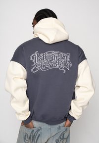 Capuchontrui met een grijze body en crèmekleurige mouwen, met een groot decoratief logo op de achterkant in wit. Zachte, gestructureerde stof.