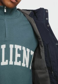 Pullover lavorato a maglia color teal con la scritta "RESILIENT" in bianco sul davanti, abbinato a un giubbotto imbottito nero con tasche con zip e una fodera grigia.