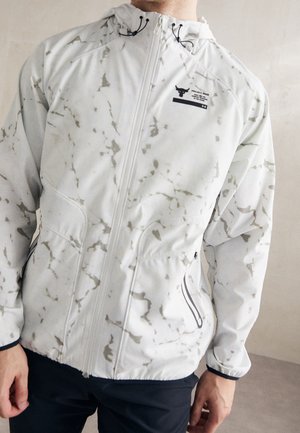Weißes Jackett mit grauem Marmor-Muster, Kapuze und schwarzem Logo auf der Brust, getragen von einer Person vor neutralem Hintergrund.