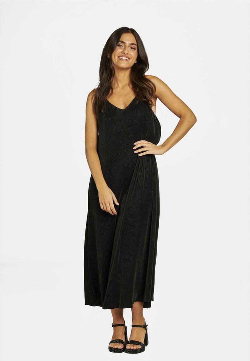 Robe maxi noire avec un décolleté en V, un tissu texturé et un design drapé. Présente une coupe loose et une silhouette jusqu'aux chevilles. Chaussures noires à lanières.