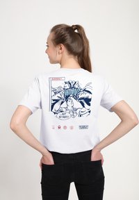 Bílý bavlněný tričko s grafickým motivem ve stylu komiksu na zadní straně v námořnické modři, zobrazující postavy a text v obdélníkovém designu.