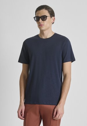 Giovane uomo con occhiali da sole neri, maglietta a maniche corte blu navy e pantaloni color ruggine su sfondo grigio uniforme.