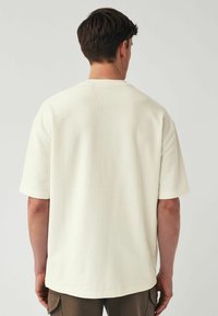 T-shirt in cotone crema con vestibilità rilassata, maniche corte e tessuto strutturato. Design semplice, orlo leggermente curvato, adatta per un abbigliamento casual.