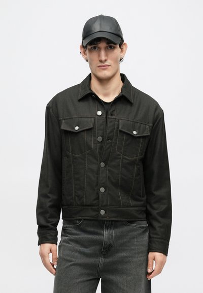 Jachetă din denim negru cu cusături vizibile, două buzunare pe piept și nasturi metalici. Pur tată cu o cămașă închisă la culoare și blugi gri. Se adaugă o șapcă neagră.