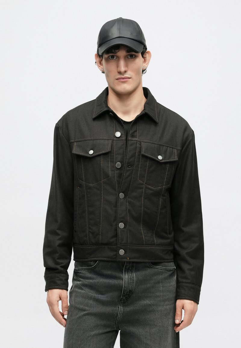 Sort denimjakke med synlig syning, to brystlommer og metaltryknapper. Bæres med en mørk skjorte og grå jeans. Sort kasket tilføjet.