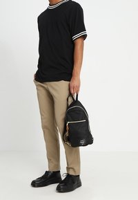 Zaino nero con zip dorate, tessuto testurizzato e un piccolo etichetta in pelle. Forma arrotondata e manico superiore per un facile trasporto.