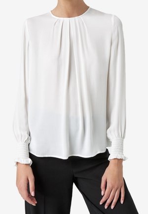 Femme portant un chemisier blanc à manches longues avec des fronces au niveau de l'encolure et des poignets smockés, assorti à un pantalon noir.