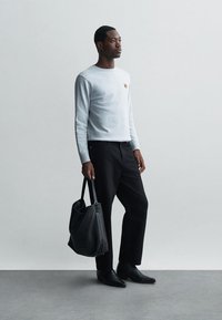 Hellgraues Langarmshirt mit Logo, schwarze Hose und schwarze Lederhandtasche. Der Mann steht in einem neutralen Raum und trägt schwarze Schuhe.
