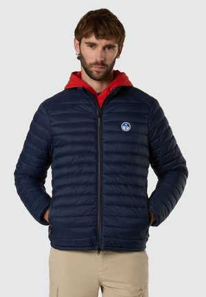 Mann mit kurzen braunen Haaren und Bart, trägt eine marineblaue gesteppte Jacke über einem roten Kapuzenpullover, Hände in den Taschen, beige Hose, neutraler grauer Hintergrund.