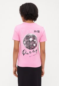 Rosa bomull T-skjorte med en stor diskokulegrafikk på baksiden, med teksten "90s VIBE" og "Party" i kontrasterende farger. Korte ermer.