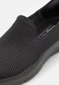 Svart mesh sko utan snörning med en texturerad ovandel, minimal sömnad och en slät, vadderad sula med en lätt kurva.