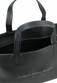 Schwarze Ledertasche mit doppelten Griffen, gefüttertem Innenraum, geprägtem "Calvin Klein Jeans"-Logo in Grün und glatter Textur.