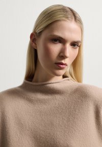 Maglione di lana beige con collo alto, caratterizzato da una texture morbida e un delicato motivo a maglia. Primo piano della parte superiore del corpo e del volto.