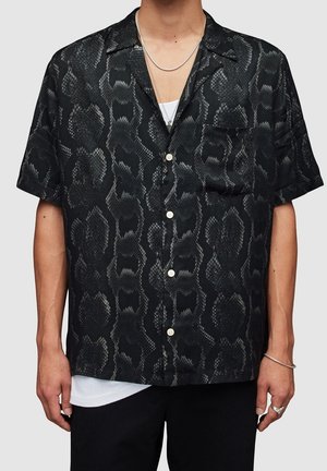 Uomo che indossa una camicia nera a maniche corte con motivo di stampa serpenteggio grigio scuro e una maglietta bianca sotto.