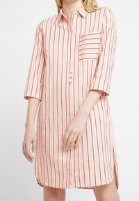 Vestido de camisa a rayas rosa y blanco con frente abotonado, cuello, mangas tres cuartos, un bolsillo en el pecho y aberturas laterales.