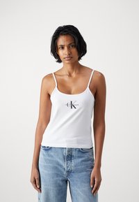 Hvitt tank topp med tynne stropper, med et trykket "Calvin Klein Jeans"-logo i mørkegrått. Laget av glatt, lett stoff.