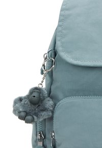 Zaino in tessuto azzurro chiaro con chiusura a patta, dotato di un portachiavi a forma di gorilla peloso grigio e tasche con cerniera. Superficie texturizzata con dettagli di cucitura.