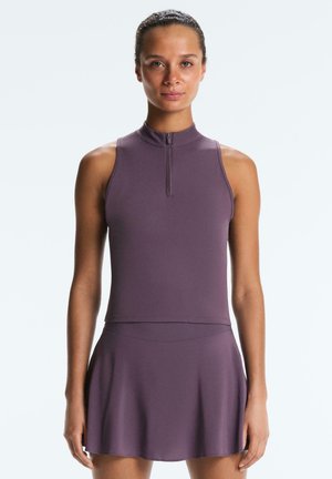 Kobieta stojąca, ubrana w bezrękawnik z fioletowym suwakiem przy szyi i pasującą rozkloszowaną spódnicę, na tle jednolitej białej ściany.