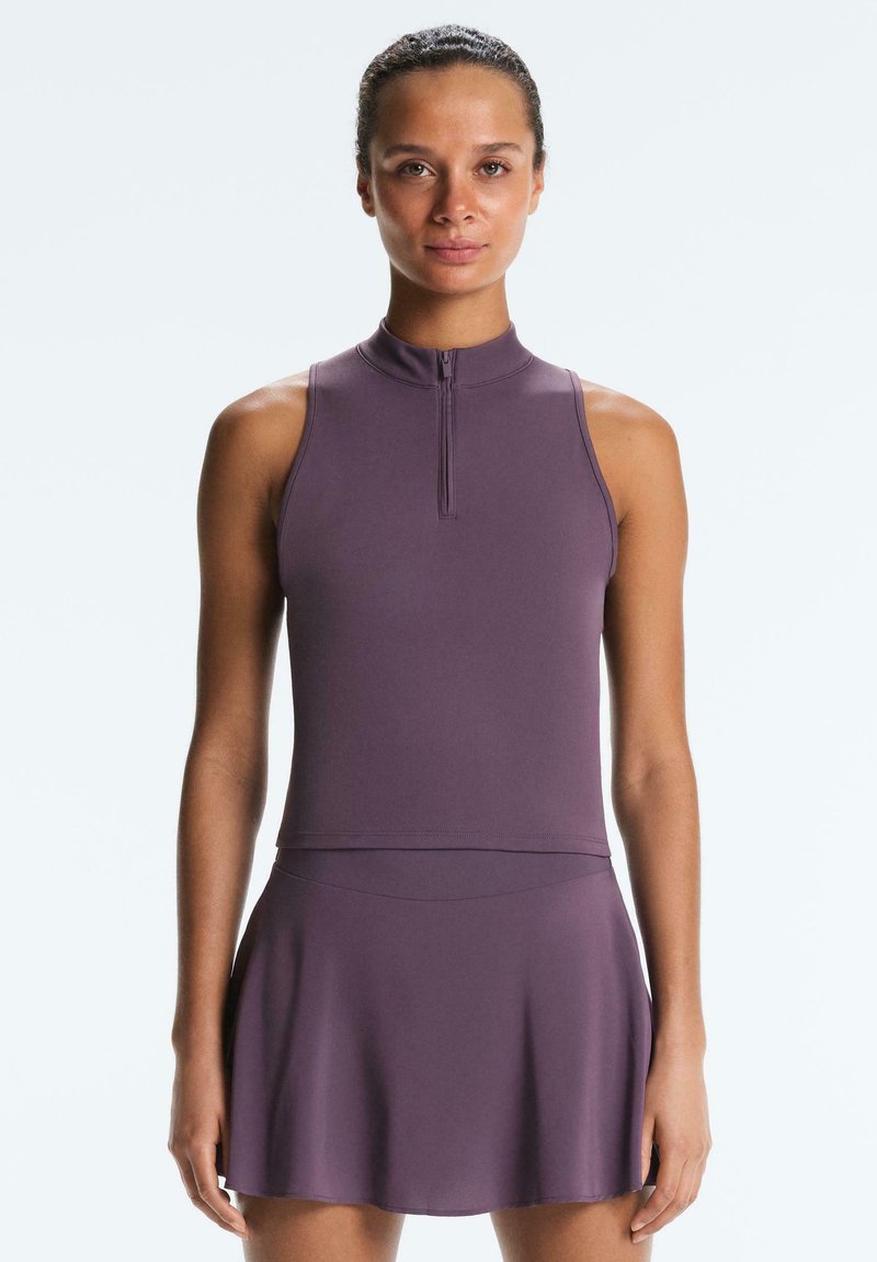 Femme debout portant un haut sans manches violet à col zippé et une jupe évasée assortie, sur un fond blanc uni.