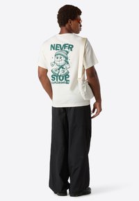 Mand iført en cremefarvet T-shirt med "Never Stop Exploring" grafik på ryggen, sorte brede bukser, orange solbriller og en hvid skuldertaske.