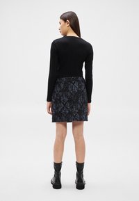 Femme debout de dos, portant un haut noir à manches longues, une jupe texturée foncée et des bottines noires, sur un fond blanc uni.