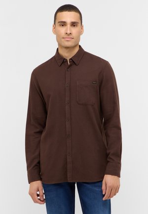 Junger Mann mit kurzen Haaren, der ein dunkelbraunes Hemd mit Brusttasche und blaue Jeans trägt, vor einem schlichten hellen Hintergrund.