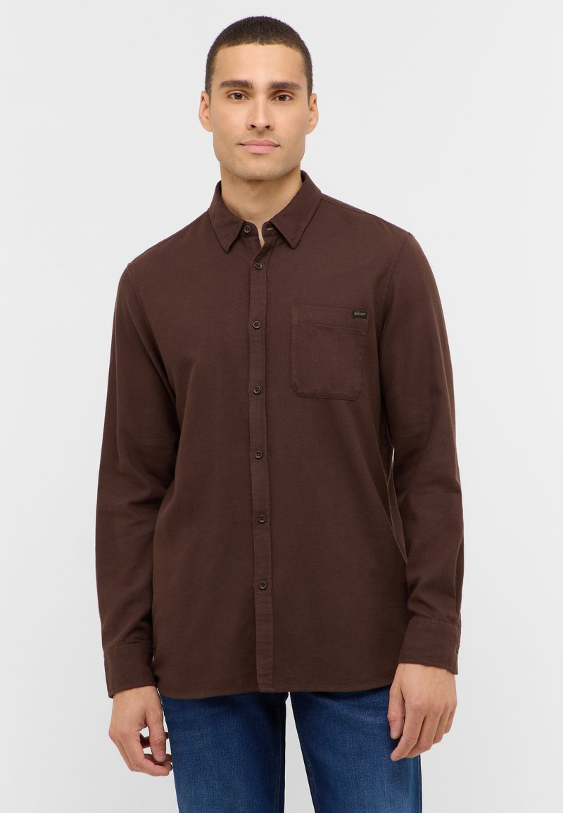 Junger Mann mit kurzen Haaren, der ein dunkelbraunes Hemd mit Brusttasche und blaue Jeans trägt, vor einem schlichten hellen Hintergrund.