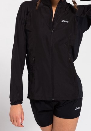 Person trägt eine schwarze Asics Zip-Up-Jacke und passende schwarze Asics-Shorts, steht mit erhobenem Arm.