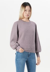 Noor naine pikkade blondide juustega, seljas lavendlitoonis puskpöördedega sviiter ja sinised teksad, poseerib üks käsi taskus.